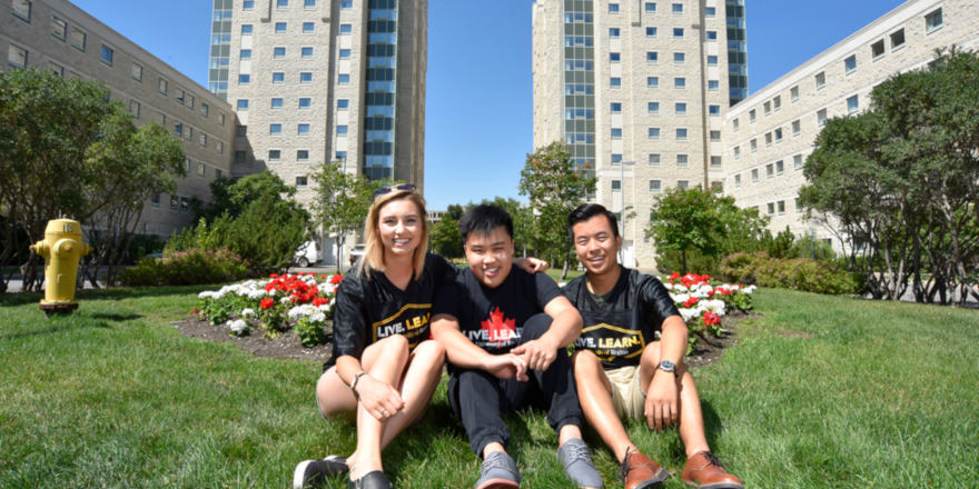 A group of students sits on the grass before the University of Regina campus, smiling.