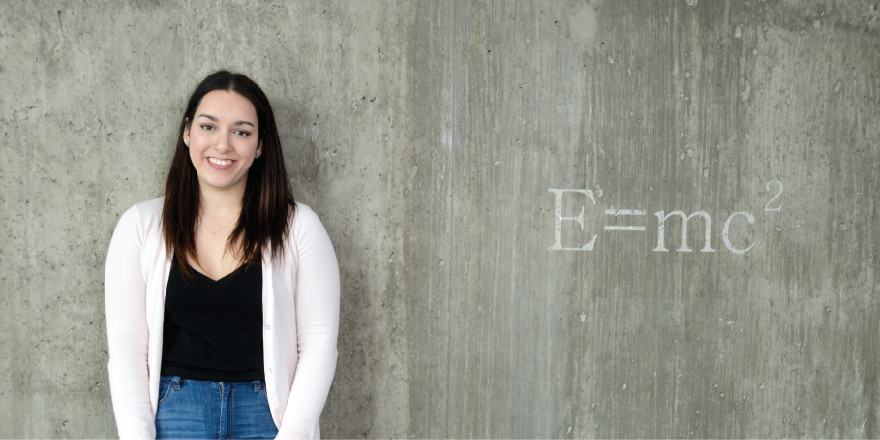 Lily, a University of Regina student, smiles, with E=mc squared on the wall beside her.
